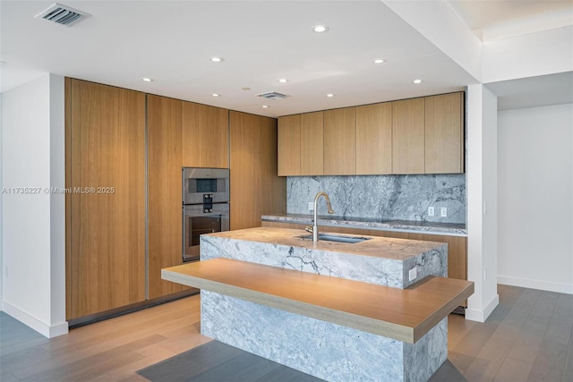 kitchen with sink, tasteful backsplash, light hardwood / wood-style flooring, an island with sink, and double oven