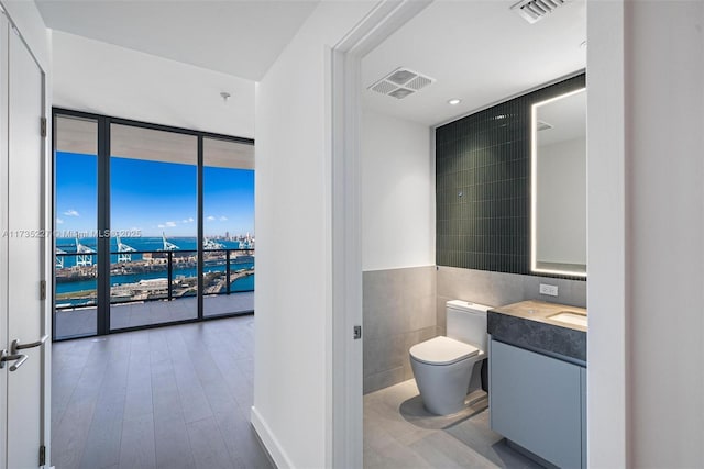 bathroom with toilet, a water view, tile walls, vanity, and a wall of windows