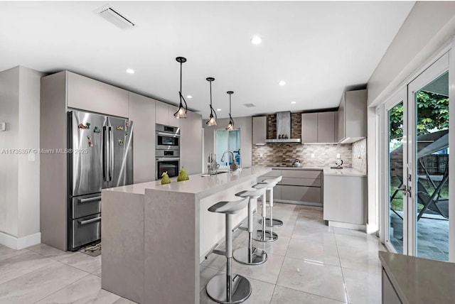 kitchen featuring pendant lighting, wall chimney range hood, appliances with stainless steel finishes, gray cabinetry, and a kitchen bar