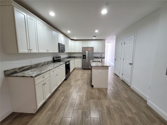 kitchen with a center island with sink, dark stone counters, stainless steel appliances, white cabinetry, and a sink