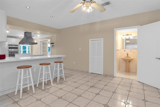 kitchen featuring a breakfast bar area, ceiling fan, white cabinetry, island exhaust hood, and kitchen peninsula