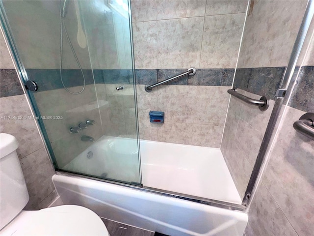 bathroom featuring toilet, bath / shower combo with glass door, and tile walls