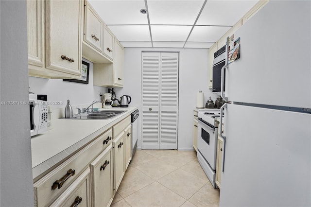 kitchen featuring sink, white appliances, cream cabinets, light tile patterned flooring, and a drop ceiling