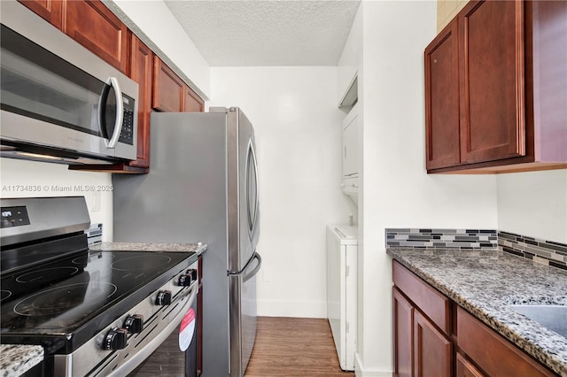 kitchen featuring appliances with stainless steel finishes, hardwood / wood-style flooring, stacked washer / dryer, light stone counters, and a textured ceiling