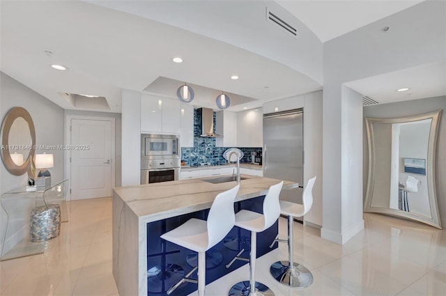 kitchen featuring wall chimney range hood, a breakfast bar area, built in appliances, white cabinets, and a center island with sink