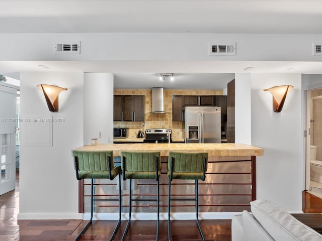 kitchen featuring dark brown cabinetry, appliances with stainless steel finishes, a kitchen bar, and wall chimney exhaust hood