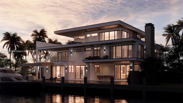 back of house at dusk featuring a water view, a balcony, and stucco siding