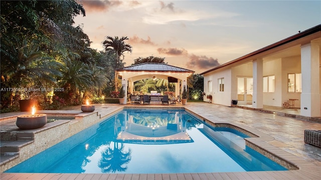 pool at dusk featuring a gazebo, an outdoor living space with a fire pit, and a patio