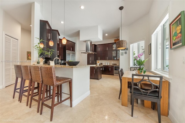 kitchen featuring light stone counters, dark brown cabinetry, hanging light fixtures, and wall chimney range hood