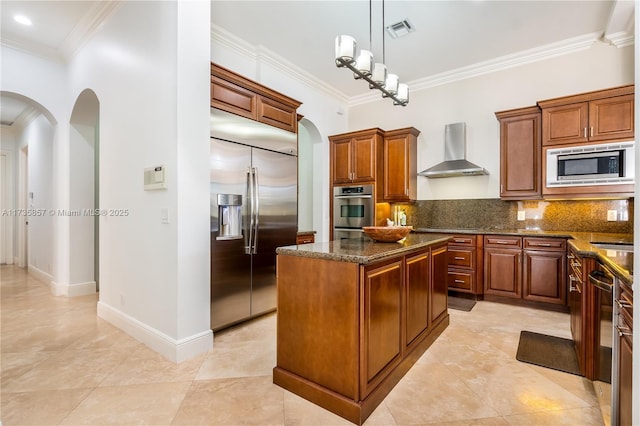 kitchen with dark stone countertops, a center island, built in appliances, decorative backsplash, and wall chimney exhaust hood