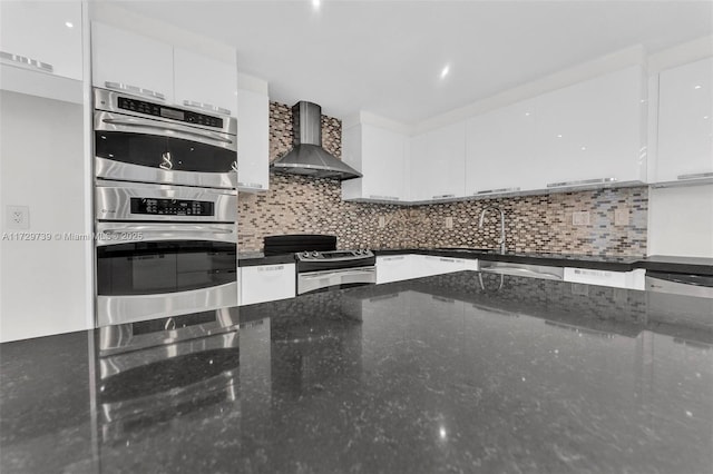 kitchen with wall chimney range hood, dark stone counters, stainless steel appliances, decorative backsplash, and white cabinets