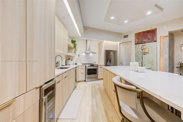 kitchen with wall chimney range hood, sink, appliances with stainless steel finishes, wine cooler, and light brown cabinetry