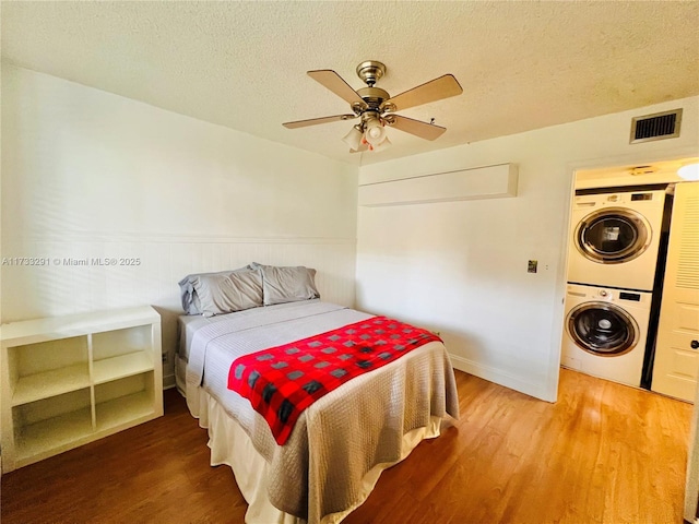 bedroom featuring wood-type flooring, stacked washer and clothes dryer, ceiling fan, and a textured ceiling