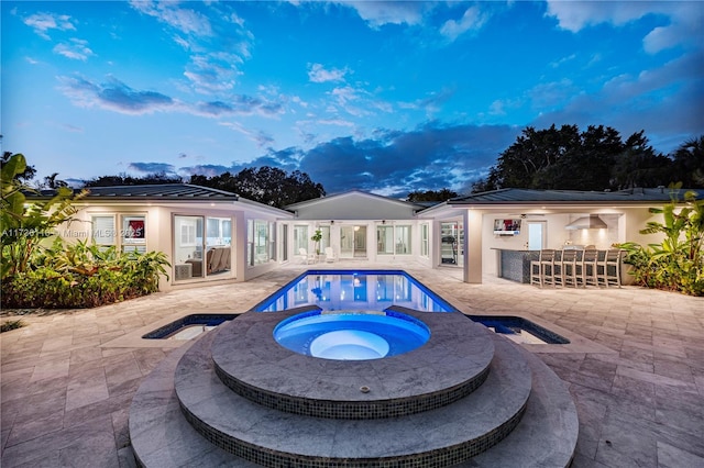 pool at dusk with an outdoor pool, a patio area, outdoor dry bar, and an in ground hot tub