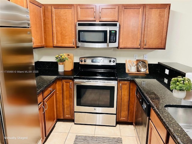 kitchen with light tile patterned floors, stainless steel appliances, dark stone counters, and brown cabinets