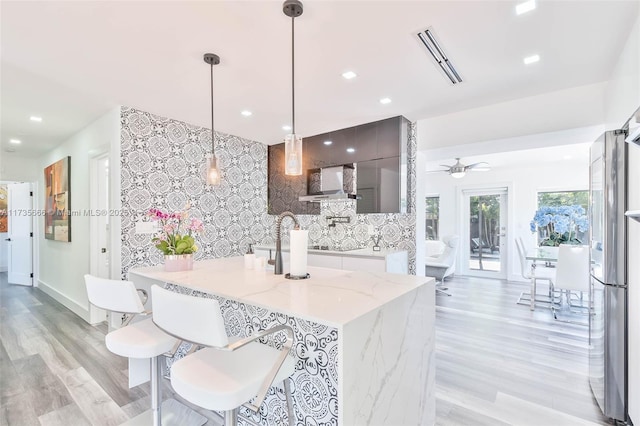 kitchen featuring hanging light fixtures, light wood-type flooring, stainless steel fridge, light stone countertops, and wall chimney range hood