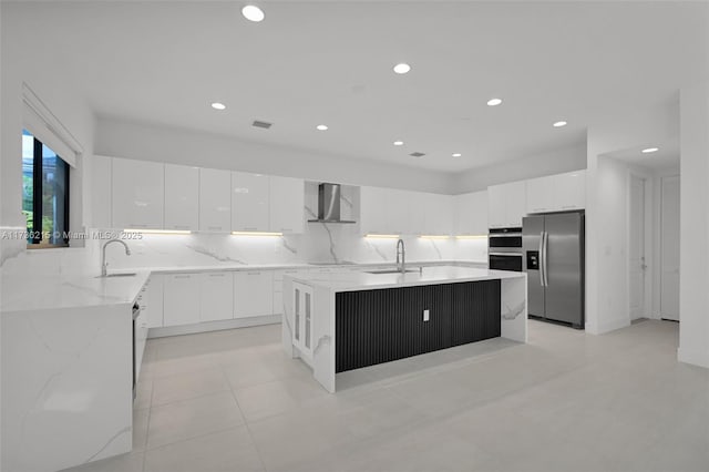 kitchen featuring wall chimney range hood, sink, white cabinetry, stainless steel appliances, and an island with sink