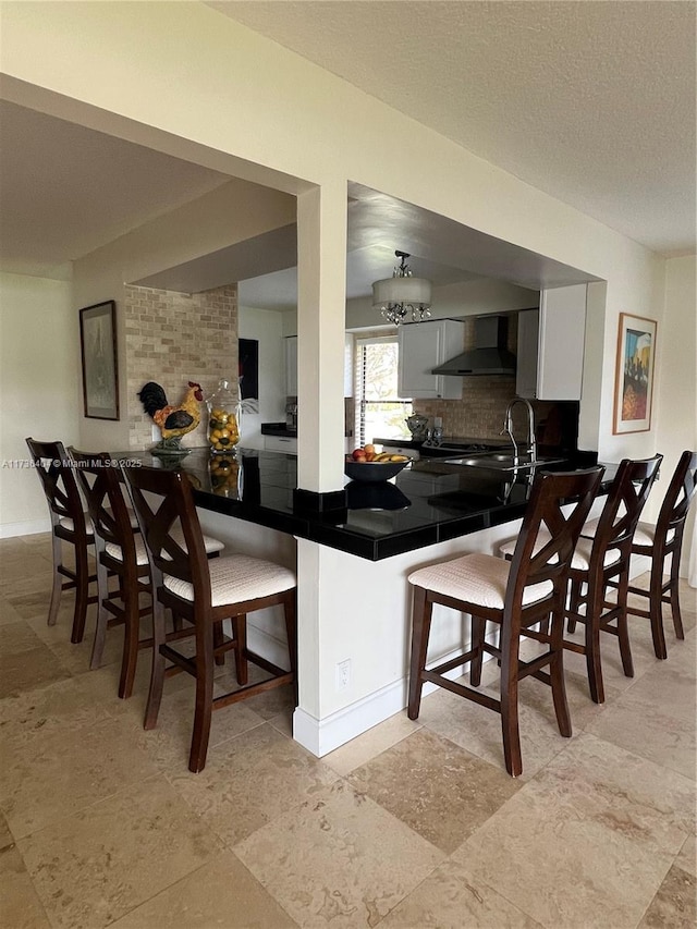 kitchen featuring tasteful backsplash, a kitchen breakfast bar, kitchen peninsula, white cabinets, and wall chimney range hood