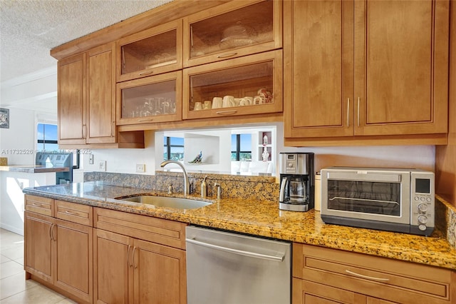 kitchen with stainless steel dishwasher, glass insert cabinets, a sink, a textured ceiling, and light stone countertops