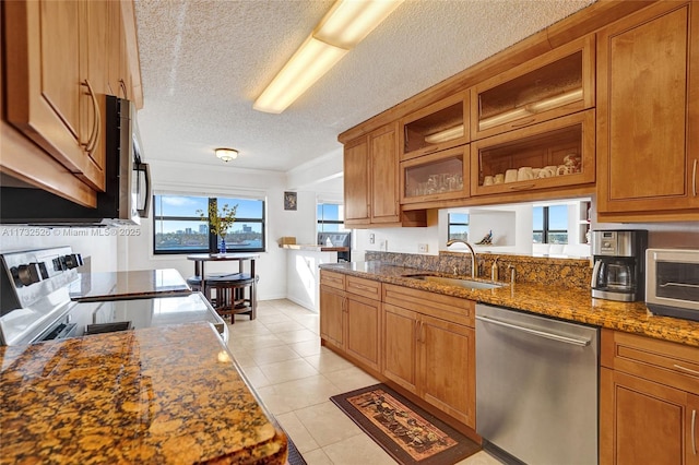 kitchen with light tile patterned floors, dark stone counters, appliances with stainless steel finishes, brown cabinets, and a sink