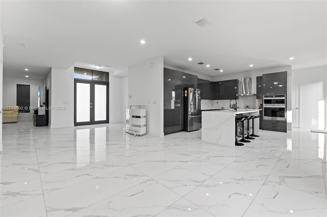 kitchen with appliances with stainless steel finishes, a breakfast bar, a kitchen island with sink, wall chimney exhaust hood, and french doors