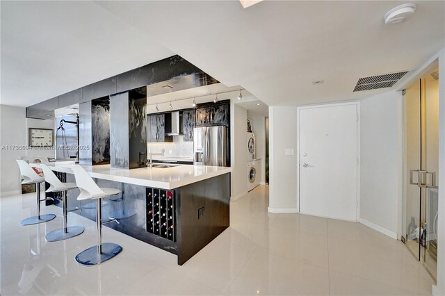 kitchen with wall chimney exhaust hood, a kitchen bar, stainless steel fridge, stacked washer / dryer, and decorative backsplash