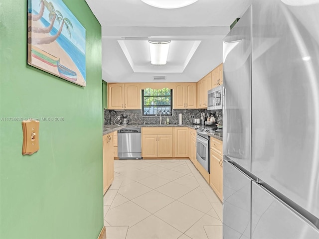 kitchen featuring appliances with stainless steel finishes, backsplash, light brown cabinetry, a tray ceiling, and dark countertops