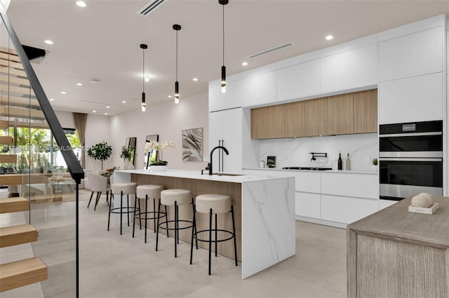 kitchen with sink, white cabinetry, a kitchen island with sink, hanging light fixtures, and decorative backsplash