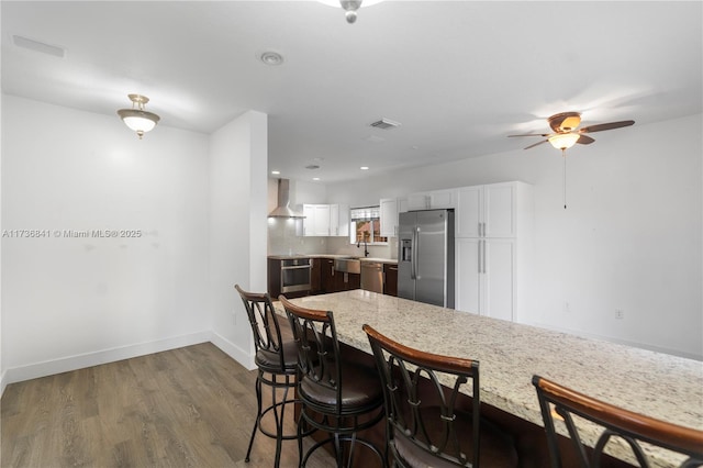 kitchen with wall chimney exhaust hood, wood-type flooring, stainless steel appliances, light stone countertops, and white cabinets