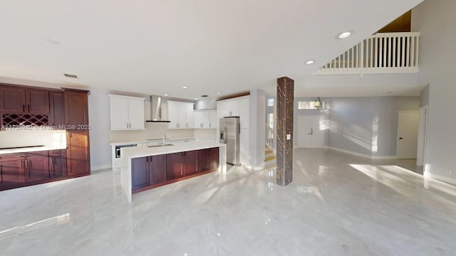 kitchen featuring wall chimney range hood, sink, white cabinetry, a center island with sink, and stainless steel fridge with ice dispenser