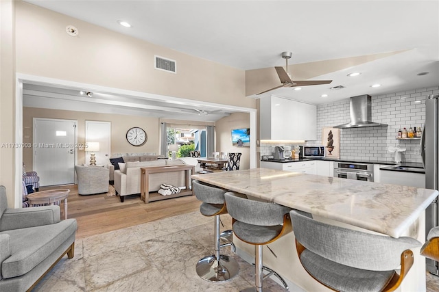 kitchen with appliances with stainless steel finishes, tasteful backsplash, white cabinets, ceiling fan, and wall chimney range hood