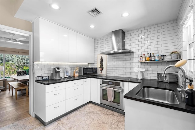 kitchen with wall chimney exhaust hood, sink, white cabinetry, appliances with stainless steel finishes, and ceiling fan