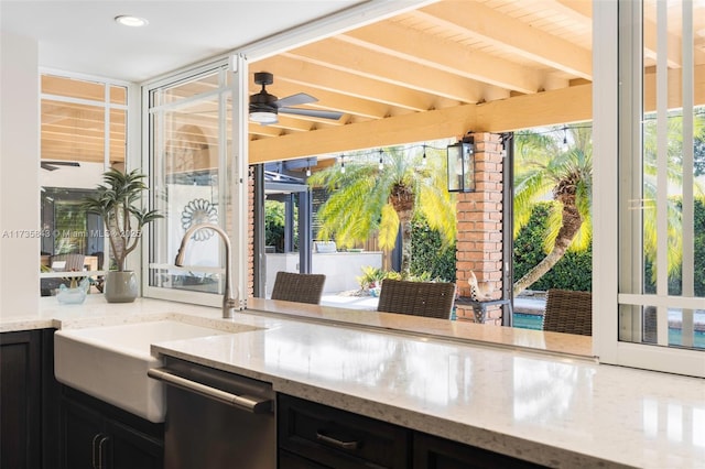 kitchen with a ceiling fan, a sink, light stone counters, dark cabinetry, and dishwasher