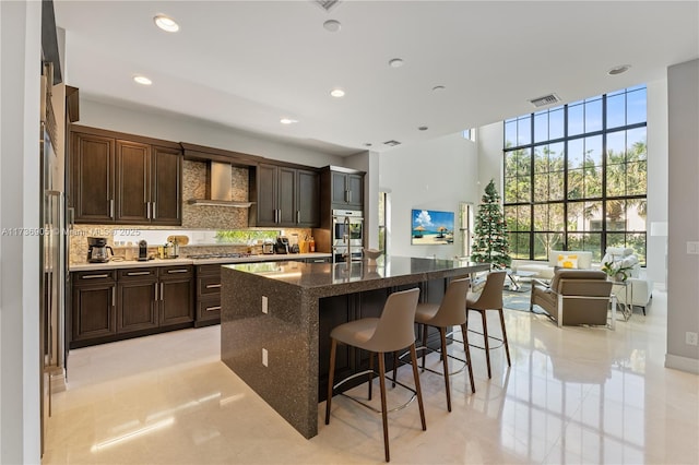 kitchen with a breakfast bar, backsplash, stainless steel appliances, an island with sink, and wall chimney exhaust hood