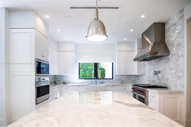 kitchen with light stone counters, decorative light fixtures, wall chimney range hood, stainless steel appliances, and white cabinets