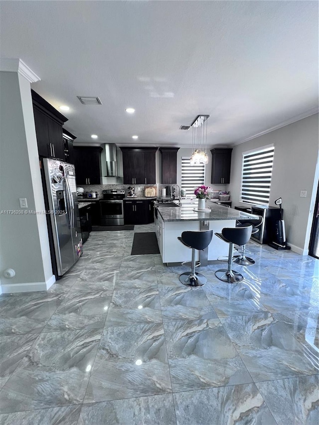 kitchen featuring wall chimney range hood, appliances with stainless steel finishes, a kitchen island with sink, ornamental molding, and decorative light fixtures