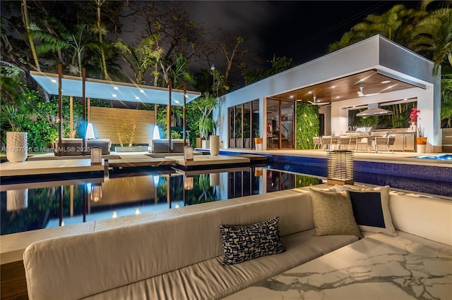 pool at night with a patio area, outdoor lounge area, ceiling fan, and an outdoor kitchen