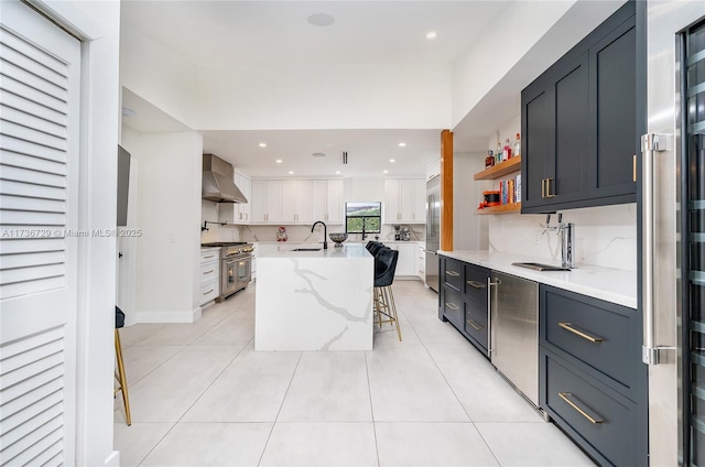 kitchen featuring wall chimney exhaust hood, a kitchen bar, high end appliances, a kitchen island with sink, and white cabinets