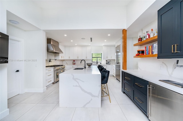 kitchen with wall chimney range hood, sink, blue cabinetry, premium appliances, and white cabinets