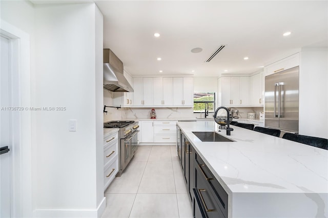 kitchen featuring wall chimney range hood, sink, white cabinetry, premium appliances, and light stone countertops