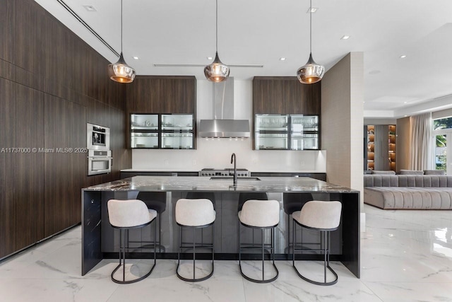 kitchen with pendant lighting, dark stone counters, a kitchen island with sink, stainless steel oven, and wall chimney range hood