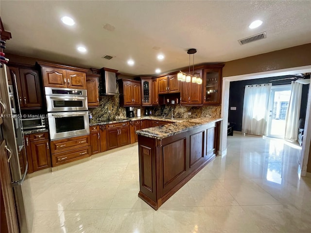 kitchen with stainless steel double oven, decorative light fixtures, visible vents, wall chimney range hood, and glass insert cabinets