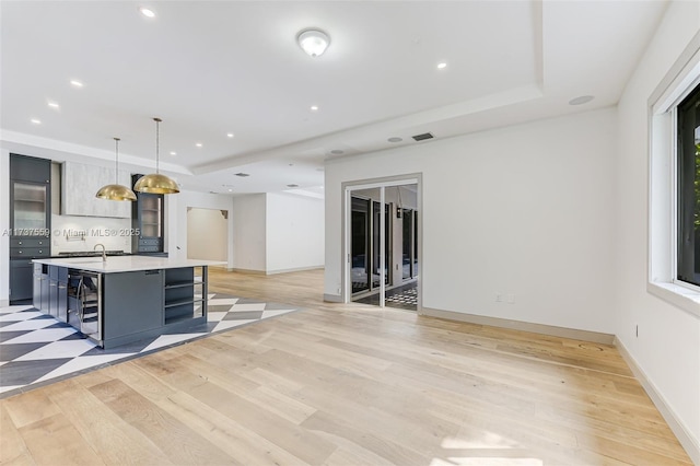 kitchen featuring a raised ceiling, a center island with sink, decorative light fixtures, beverage cooler, and light wood-type flooring