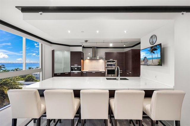 kitchen featuring sink, a kitchen breakfast bar, kitchen peninsula, dark brown cabinets, and wall chimney exhaust hood