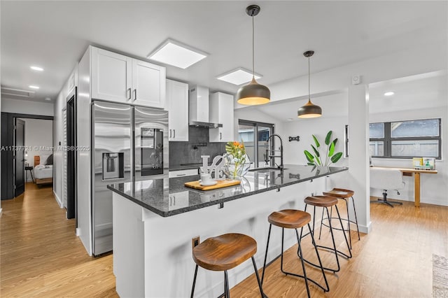 kitchen featuring wall chimney range hood, sink, white cabinetry, hanging light fixtures, and dark stone countertops