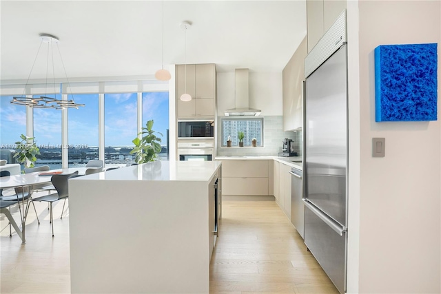 kitchen with a kitchen island, pendant lighting, built in appliances, wall chimney range hood, and light hardwood / wood-style flooring