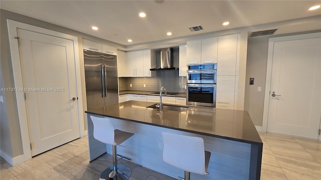 kitchen featuring wall chimney exhaust hood, sink, stainless steel appliances, a kitchen island with sink, and white cabinets