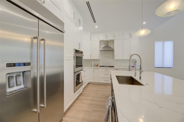 kitchen with a sink, light stone countertops, appliances with stainless steel finishes, wall chimney range hood, and white cabinets