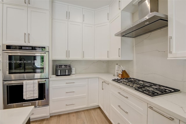 kitchen with wall chimney exhaust hood, light wood-type flooring, stainless steel appliances, decorative backsplash, and white cabinets