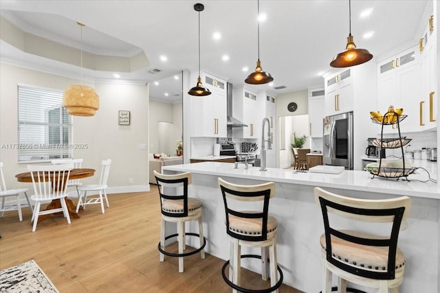 kitchen featuring appliances with stainless steel finishes, decorative light fixtures, white cabinetry, a tray ceiling, and wall chimney range hood